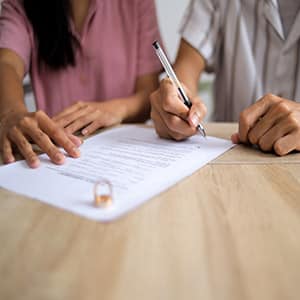 A couple signing divorce papers at a table with wedding rings in the foreground.